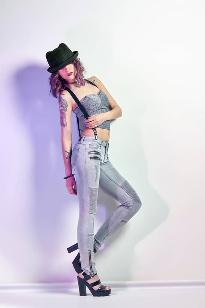 Sexy girl with purple hair and a tattoo on the body posing on grey background. Perfect woman in gray jeans and a t-shirt, bright beautiful makeup. - Foto, Bild