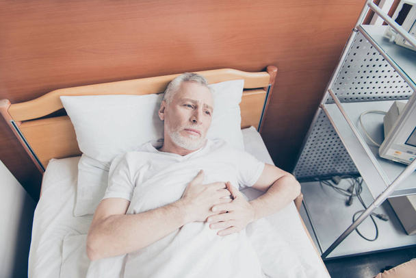Aged patient is lying in the bed and holding his hands near to h - Photo, image