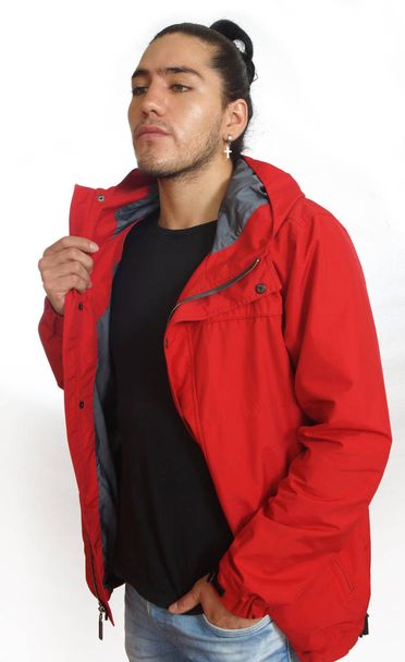 Jonge Spaanse man met verzamelde haar boog dragen zwart t-shirt en rode jas, met een hand op zijn broekzak, gedaan op zoek links - Foto, afbeelding