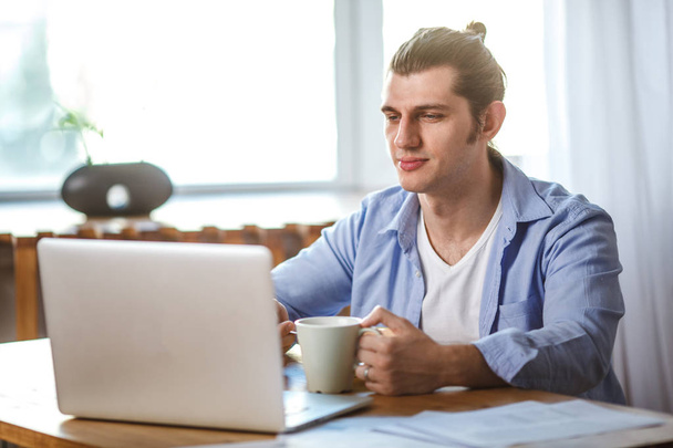 Young man holding mug, sitting at the table and looking to the laptop screen - Φωτογραφία, εικόνα