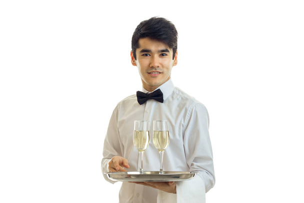 attractive young waiter in a white shirt looks straight smiles and holds on to a tray of wine glasses - Photo, Image