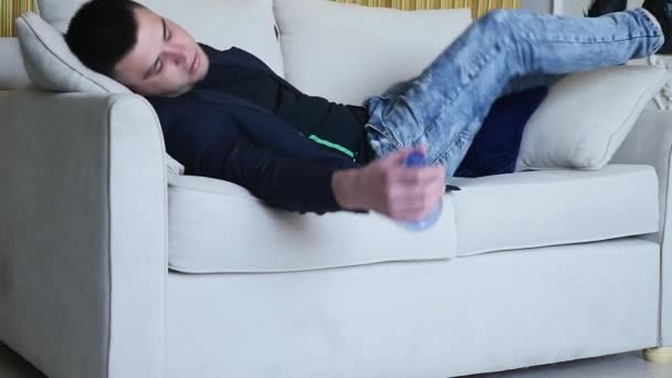 Drunk man seating on sofa . Young drunk man seating in living room with bottle in his hand on sofa, footage is taken in slow motion - Πλάνα, βίντεο