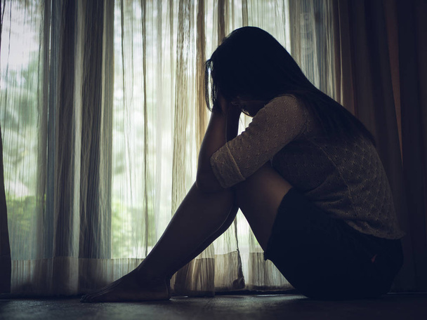 schwarz-weiße traurige Frau umarmt ihr Knie und weint. traurige Frau, die allein in einem leeren Raum neben Fenster oder Tür sitzt - Foto, Bild