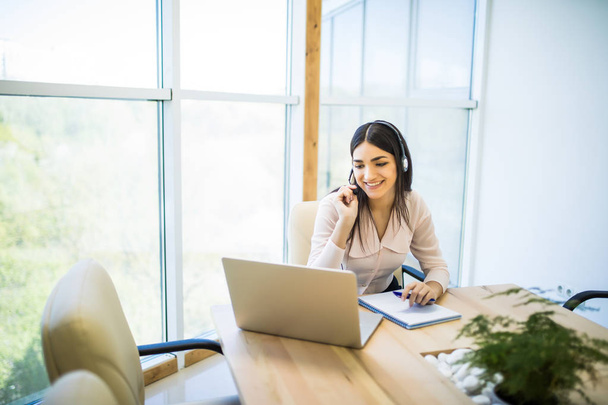 Счастливая очаровательная молодая женщина, сидящая и работающая с ноутбуком, используя гарнитуру в офисе - Фото, изображение