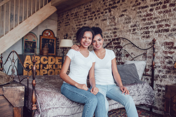 Two attractive african american girls in white T-shirts are sitting on the bed. - Foto, imagen