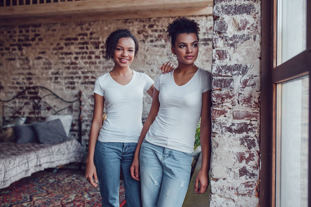 Two attractive african american girls in white T-shirts are standing near the window. - Foto, immagini