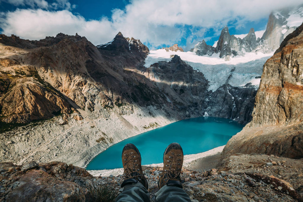 Sapatos de botas de rastreamento em um fundo azul lago entre montanhas e céu nublado
 - Foto, Imagem