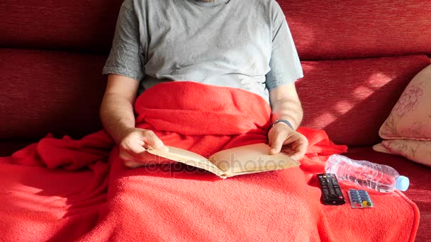 Hombre enfermo leyendo en un sillón de su casa tomar una píldora y conecta la televisión con el control a distancia
 - Metraje, vídeo