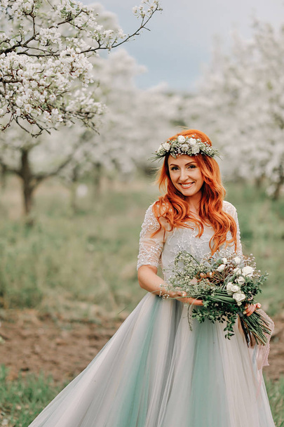Young woman in a luxurious dress is standing and smiling in a blooming garden - Foto, afbeelding