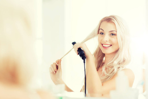 woman with styling iron doing her hair at bathroom - Photo, Image
