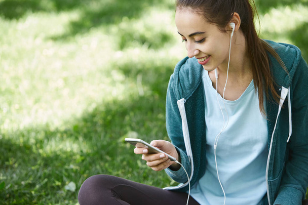 Portrait of young and sporty woman in sportswear sitting with the smartphone on the grass in park - Photo, image