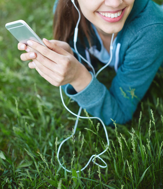 Portrait of young and sporty woman in sportswear Lies with the smartphone on the grass in park - Photo, image
