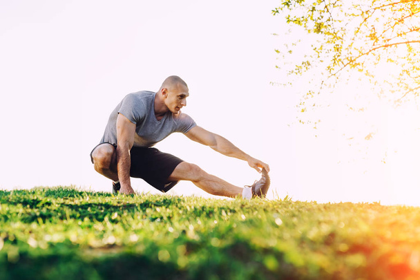 молодой спортсмен делает упражнения на растяжку, готовится к тренировке в парке. закат
 - Фото, изображение
