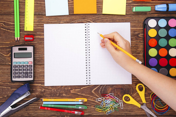 Opened blank notebook, hand with a brushwith frame of school supplies over a wooden desk background. With copy space Back to school concept. - Photo, Image