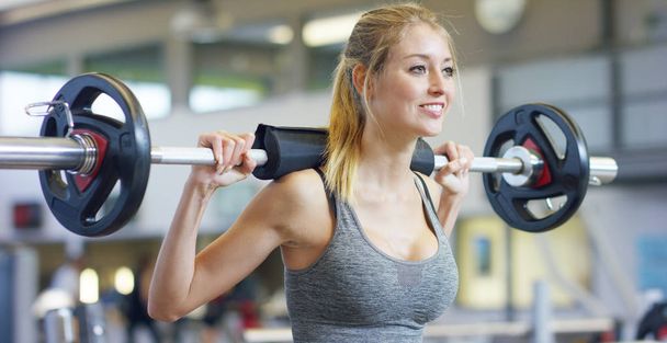 Young beautiful girl in the gym doing exercises on the squat with a barbell, improving the muscles of the buttocks and legs Concept love sports attend a gym proper nutrition slender body to be healthy - 写真・画像