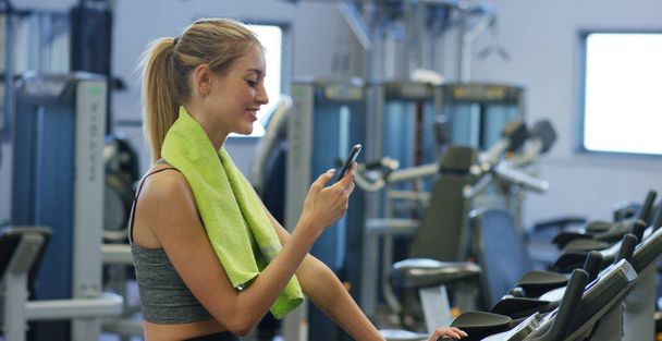 Young beautiful girl in a gym, shakes her legs on a cycling simulator, with a phone in her hand. The concept: to love sports, to attend a gym, proper nutrition, a slender body, to be healthy. - Фото, зображення