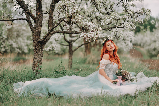 Mujer joven en un vestido de lujo se sienta en la hierba en un jardín floreciente
 - Foto, imagen