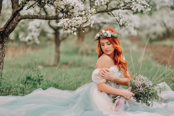 Young woman in a luxurious dress sits on the grass in a blooming garden - Photo, Image
