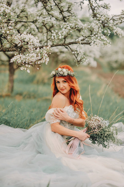 Young woman in a luxurious dress sits on the grass in a blooming garden - Foto, afbeelding
