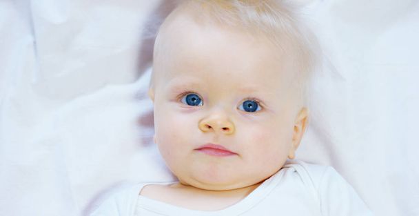 Um bebê, um menino com grandes olhos azuis e cabelos claros, senta-se e sorri em um cobertor branco de neve, olha para sua mãe, em um fundo branco
. - Foto, Imagem