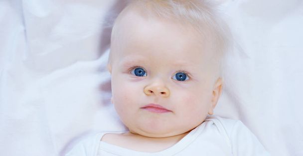 A baby, a boy with large blue eyes and light-colored hair, sits and smiles on a snow-white blanket, looks at her mother, on a white background. - 写真・画像