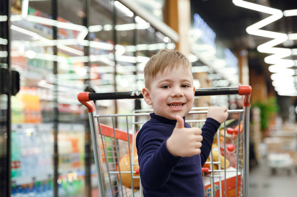 Happy boy near shopping trolley showing thumb up  - Foto, Imagen