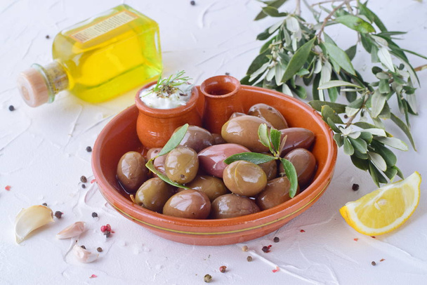 Olives in a traditional brown ceramic bowl, with tzatziki sauce, lemon, garlic, olive branch and olive oil on a white abstract background. - Foto, Bild