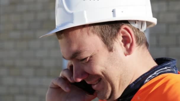 Caucasian man in helmet and vest smiling talking by phone at work place outdoor - Imágenes, Vídeo