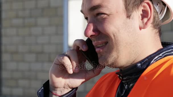 Caucasian man in helmet and vest smiling talking by phone at work place outdoor - Filmati, video
