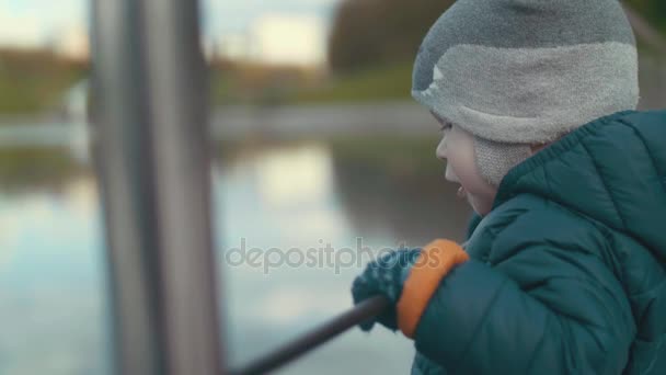 SLOW MOTION Ritratto di un grazioso bambino felice in piedi su un ponte, che si tiene per mano con corrimano, guardando uno stagno nel parco
 - Filmati, video