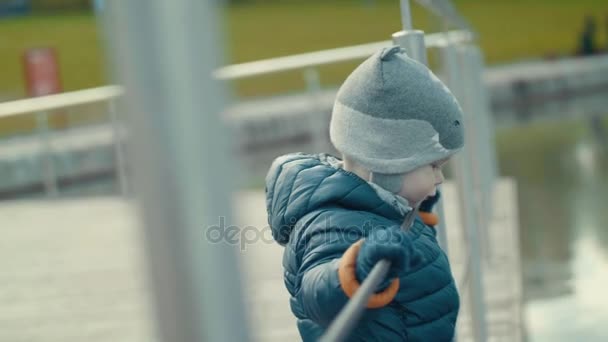 Little cute dziecko cieszy się spacer, stojącego na nabrzeżu, patrząc na staw w chłodnej pogodzie zwolnionym tempie - Materiał filmowy, wideo