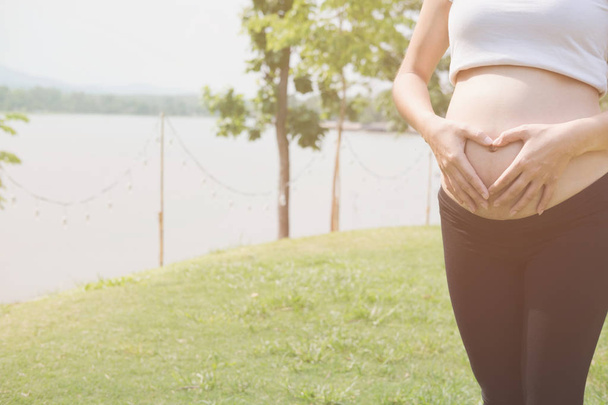 молодая беременная женщина делает знак формы сердца на животе в гаре
 - Фото, изображение