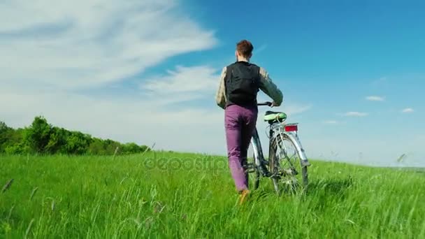 Back view: A young hipster man comes with a bicycle over a green meadow. Enjoys a walk in a picturesque rural location against the blue sky. Healthy lifestyle. Copyspace composition - Footage, Video