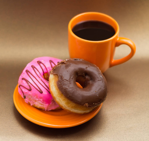 Delicious donut with pink and chocolate glazed over an orange plate, with an orange cup of coffee in a soft brown background - Photo, image