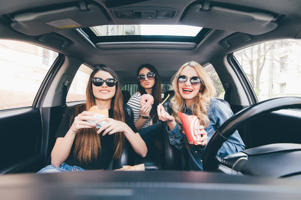 Profitant de leur déjeuner dans la voiture. Quatre belles jeunes femmes gaies se regardant avec le sourire et mangeant sortent de la nourriture tout en étant assis dans la voiture
 - Photo, image