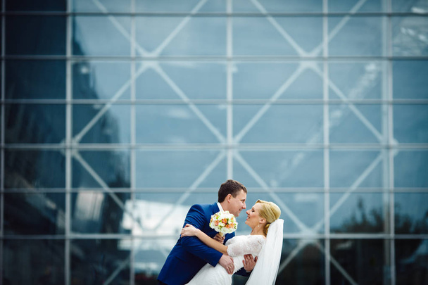 Happy wedding couple poses in the front of blue glass building - Fotoğraf, Görsel