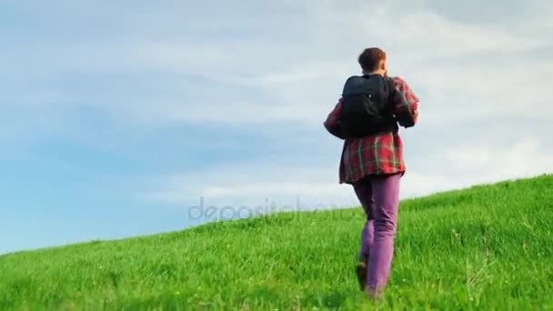 A young man with a backpack behind him rises to the top of a large green hill. In a beautiful place against the blue sky - Záběry, video