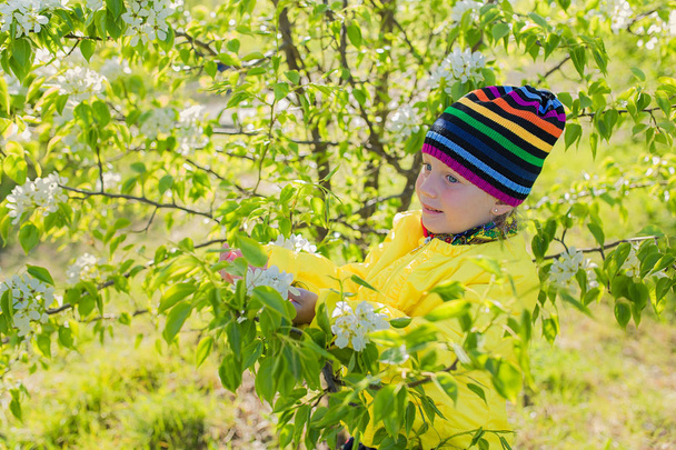 Lovely beautiful girl in a colorful hat and yellow jacket against a blooming apple tree in the spring. - Fotografie, Obrázek