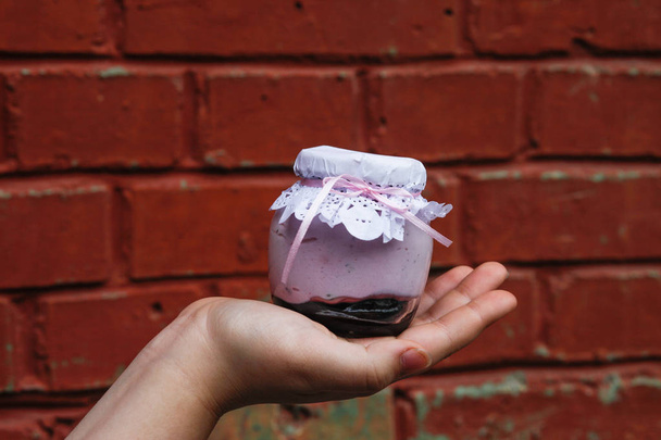 Сливочный десерт в небольшой банке глас с украшением и лентой, которая держалась в руке на деревянном фоне
 - Фото, изображение
