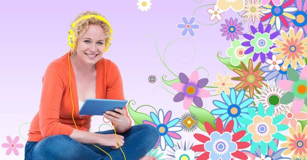 Smiling woman listening to music on headphones using tablet PC against floral background - Foto, imagen