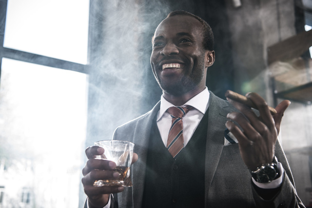 Smiling african american businessman holding glass with whiskey and smoking cigar - Fotoğraf, Görsel