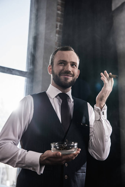 Portrait of handsome smiling bearded man holding ashtray and smoking cigar indoors - Foto, imagen