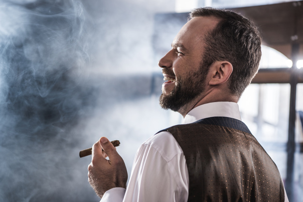 Side view of smiling confident man smoking cigar indoors  - Foto, afbeelding