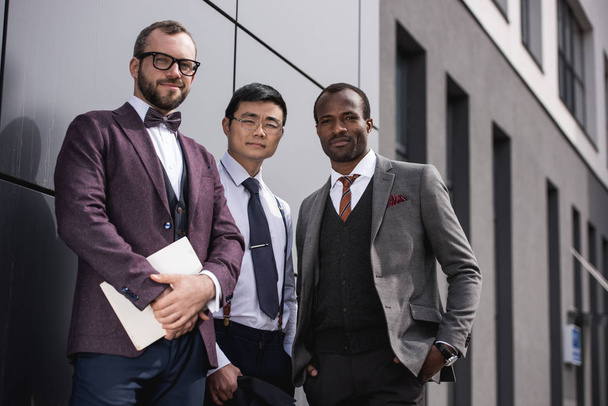 jóvenes y elegantes hombres de negocios multiétnicos en ropa formal posando al aire libre, reunión de equipo de negocios
 - Foto, Imagen