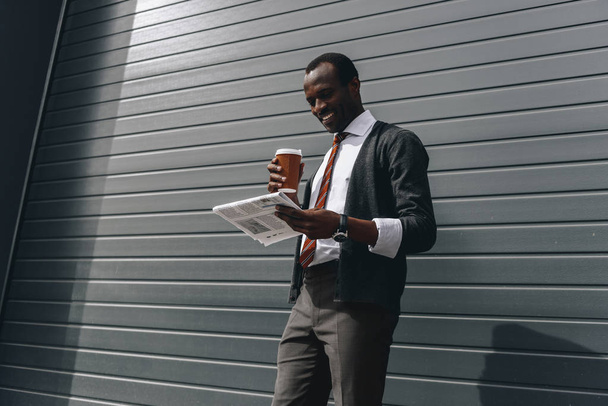 stylish african american businessman reading newspaper and holding coffee to go outdoors  - 写真・画像