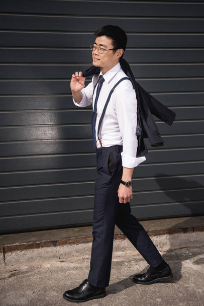 молодой улыбающийся азиатский бизнесмен в очках прогуливаясь по улице
 - Фото, изображение