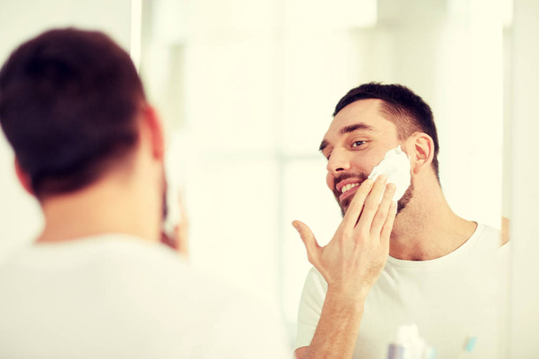 happy man applying shaving foam at bathroom mirror - Foto, afbeelding