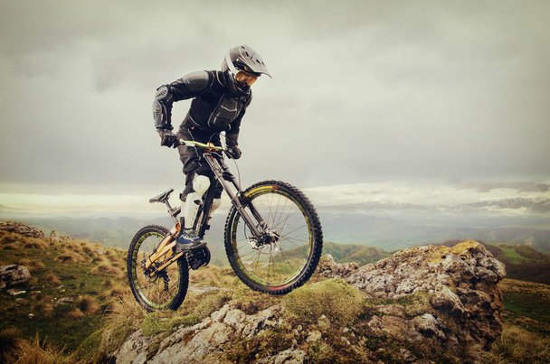 Ryder in full protective equipment on the mtb bike climbs on a rock against the backdrop of a mountain range and low clouds - Photo, Image