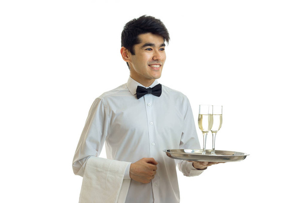 χαμογελώντας όμορφος σερβιτόρος με μαύρα μαλλιά και πουκάμισο κρατώντας ένα δίσκο με δύο ποτήρια κρασί - Φωτογραφία, εικόνα