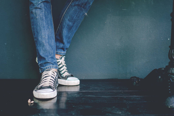 Beine eines jungen Modemachers in blauen Jeans und schwarzen Turnschuhen auf Holzboden. - Foto, Bild